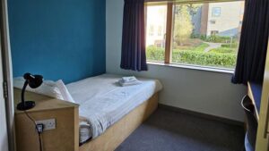 A view of a bedroom in The Hollies, showing a bed with integrated plug sockets, lamp, towels, desk and windows with curtains.