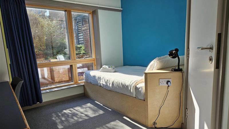 A view of a bedroom in The Hollies, showing a bed with integrated plug sockets, towels, lamp and large windows with curtains.