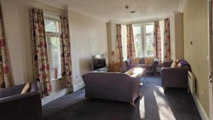 The Hollies living room area, showing a sofa and chairs spaced apart in an open room with TV, windows and curtains.
