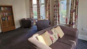 The Hollies living room area, showing a sofa, chairs and bookcase spaced apart in an open room with large windows with curtains.